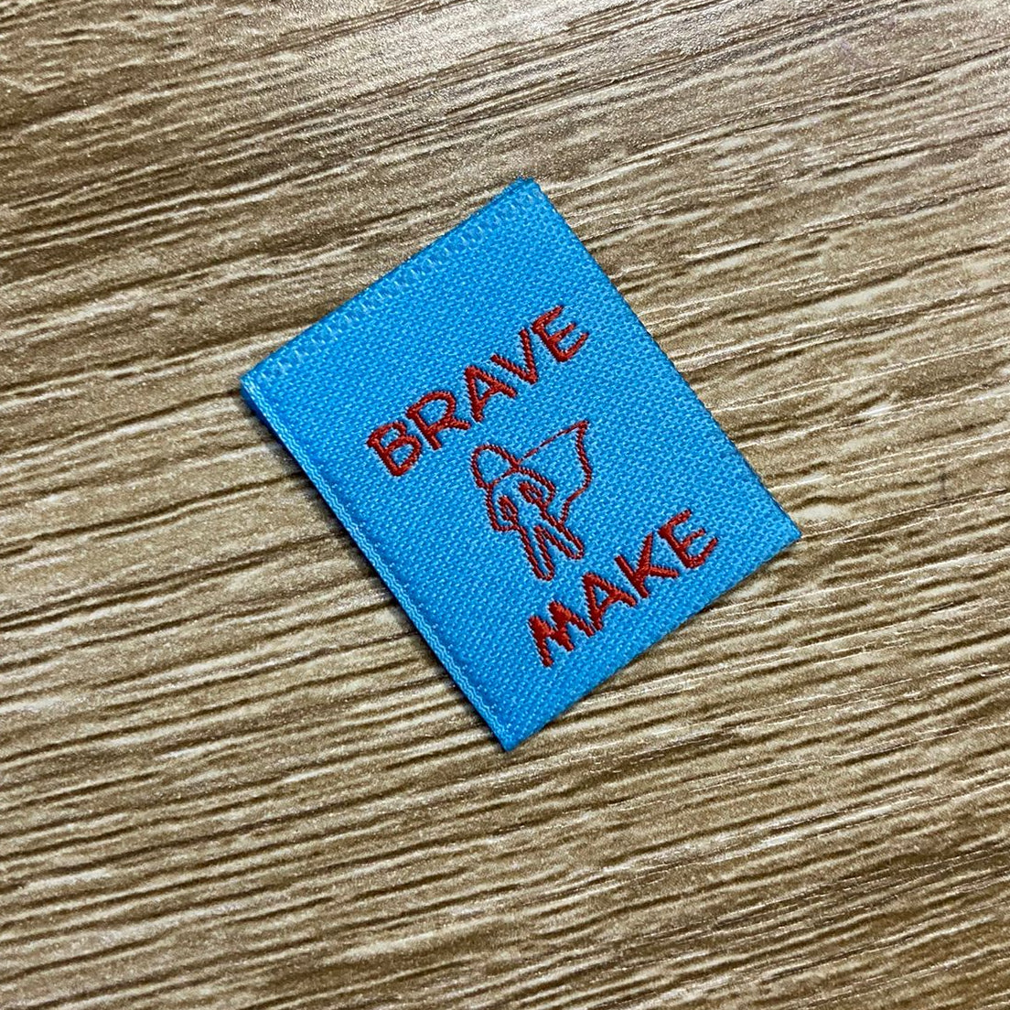 Blue woven label with 'Brave Make' text on a wooden surface