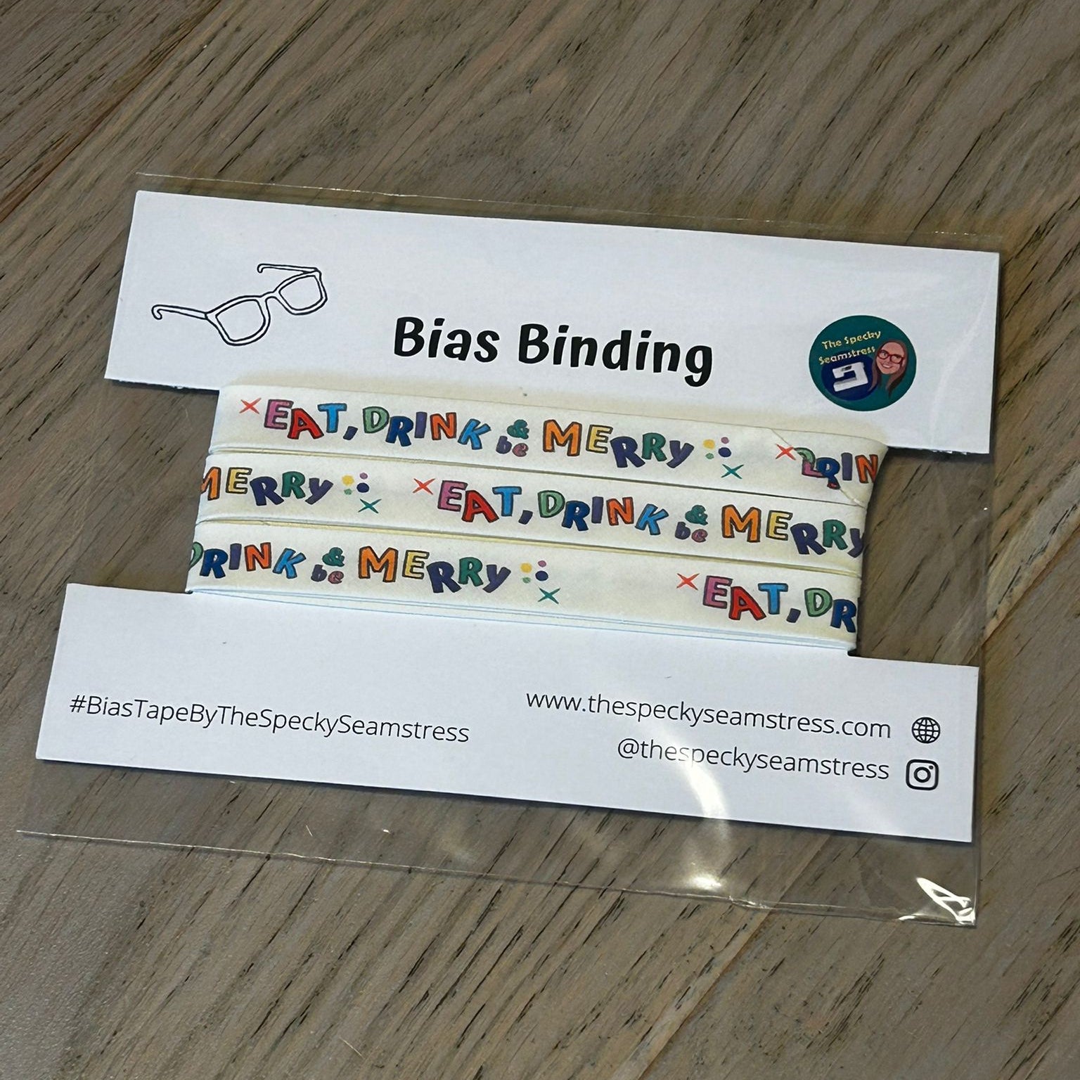 Eat, Drink and Be Merry Bias binding tape with colorful text design on a wooden surface.