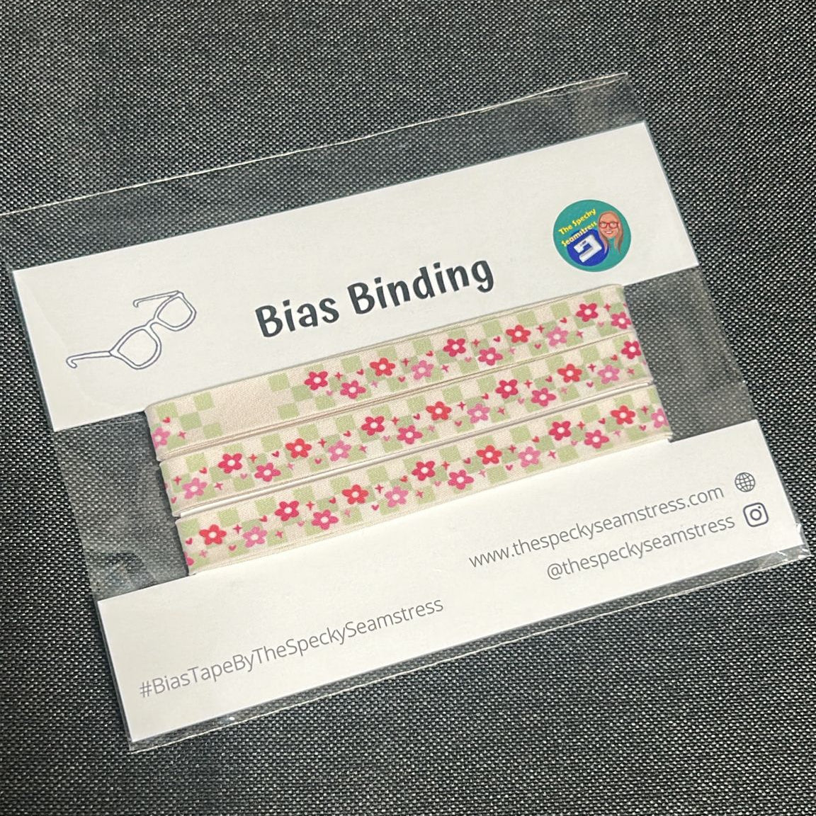 Floral Gingham Bias on a white backing card in a clear plastic package, on a grey fabric background. 