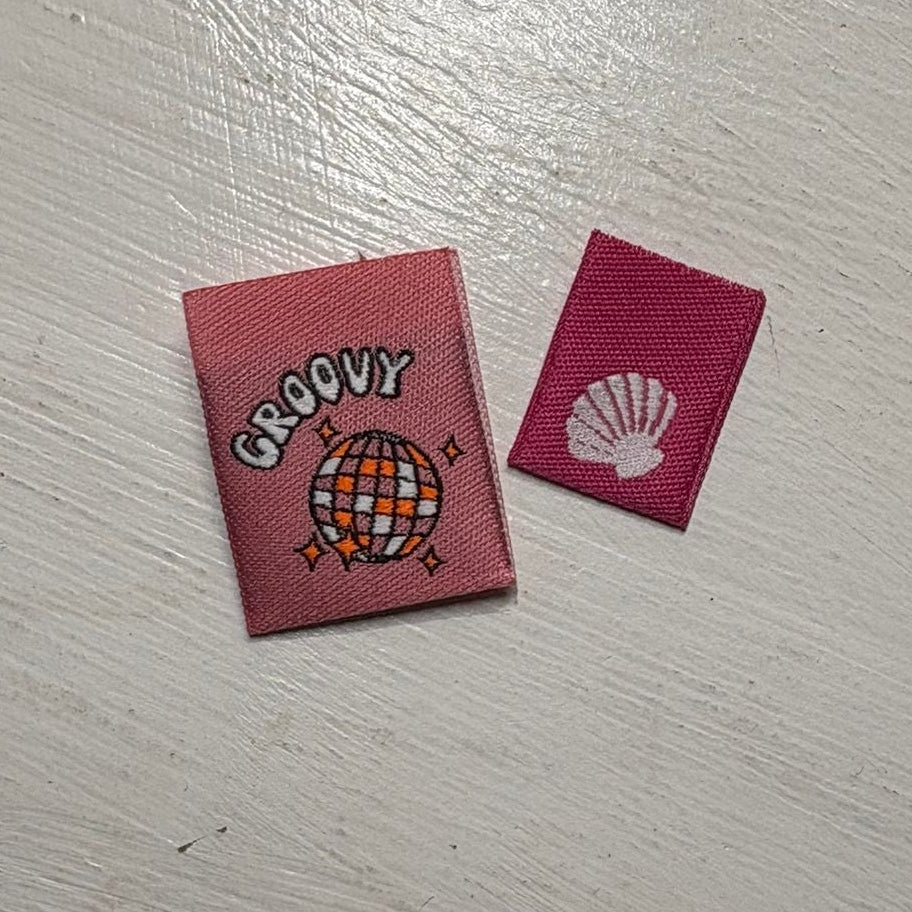 Two small pink woven labels with colourful designs on a white wooden surface. 