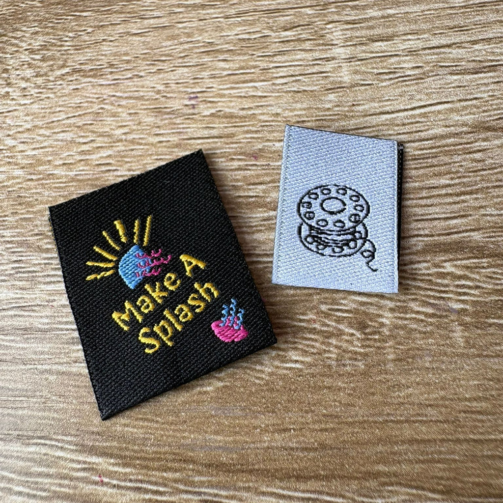 Two embroidered labels on a wooden surface, one with 'Make A Splash' text and graphics, the other with a bobbin design.