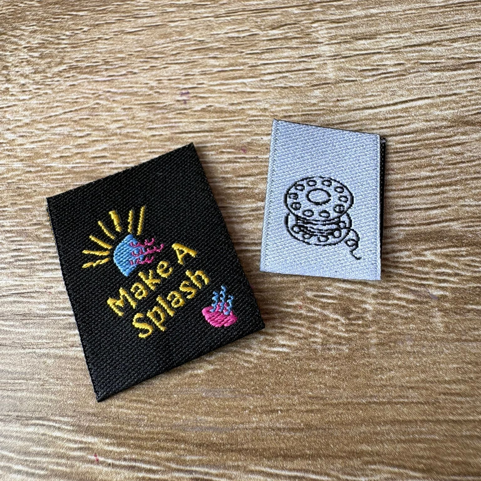 Two embroidered labels on a wooden surface, one with 'Make A Splash' text and graphics, the other with a bobbin design.
