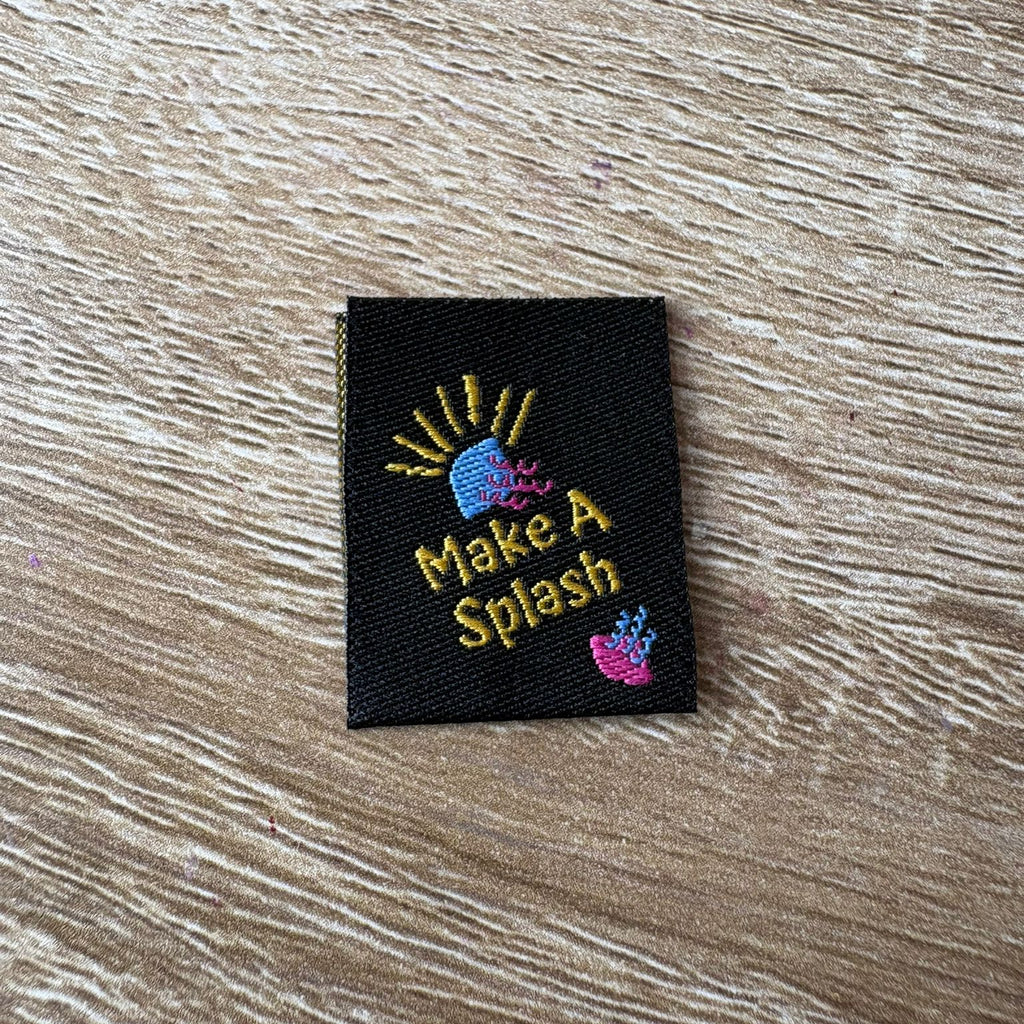 Embroidered label with 'Make A Splash' text on a wooden surface