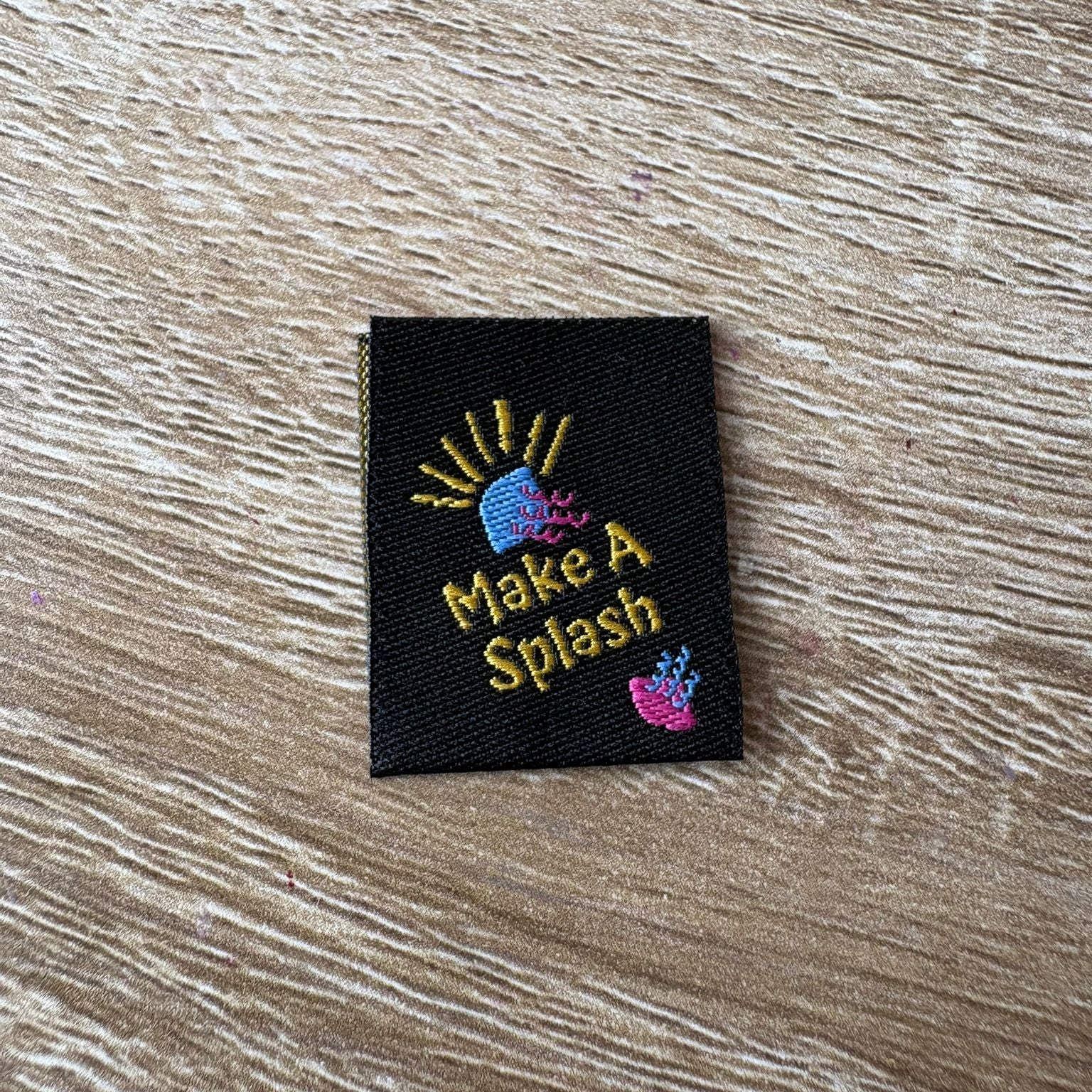 Embroidered label with 'Make A Splash' text on a wooden surface