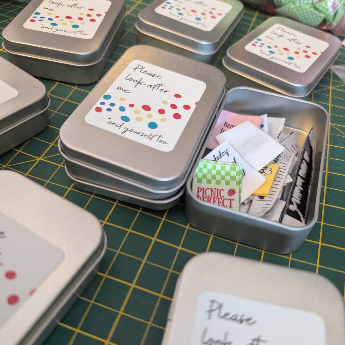 Several small silver metal tins with white labels are arranged on a green cutting mat. One tin is open and filled with small folded paper items, including a green-and-white checkered paper that reads 'PICNIC PERFECT'.