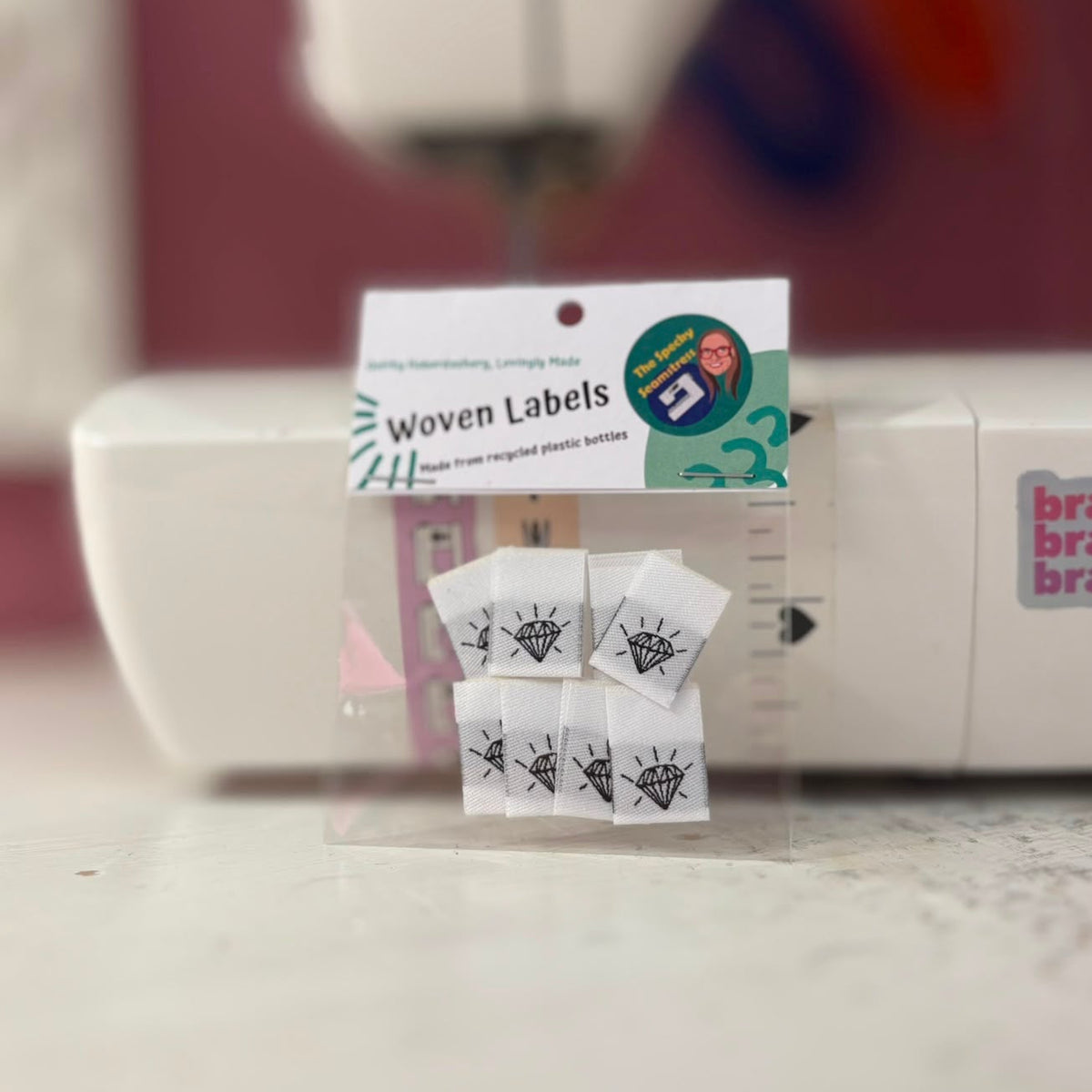 A package of 8 mini woven labels propped against a sewing machine, the white labels have a black diamond jewel outline. 