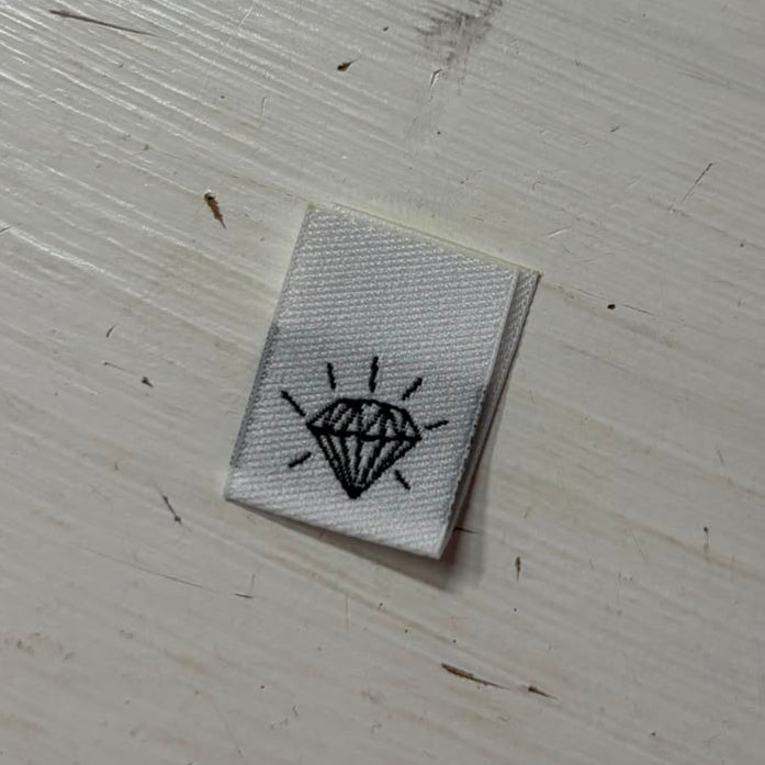 A white woven sewing label on a painted wooden surface; shows a diamond jewel outline. 