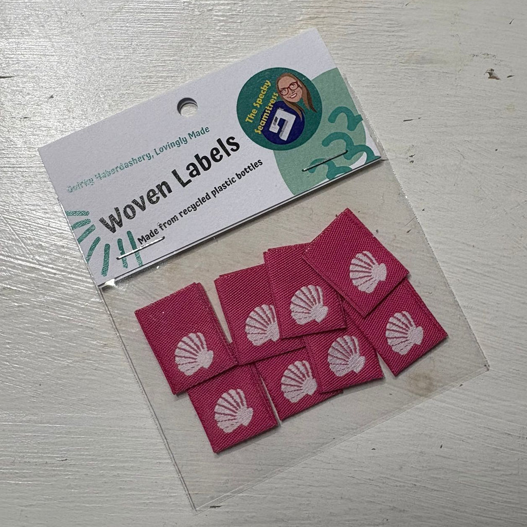 8 Woven labels with shell design in a packet on a white wooden background.