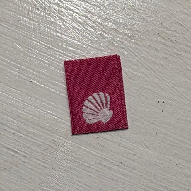 Mini hot pink woven label with a white seashell logo on a white wooden background. 