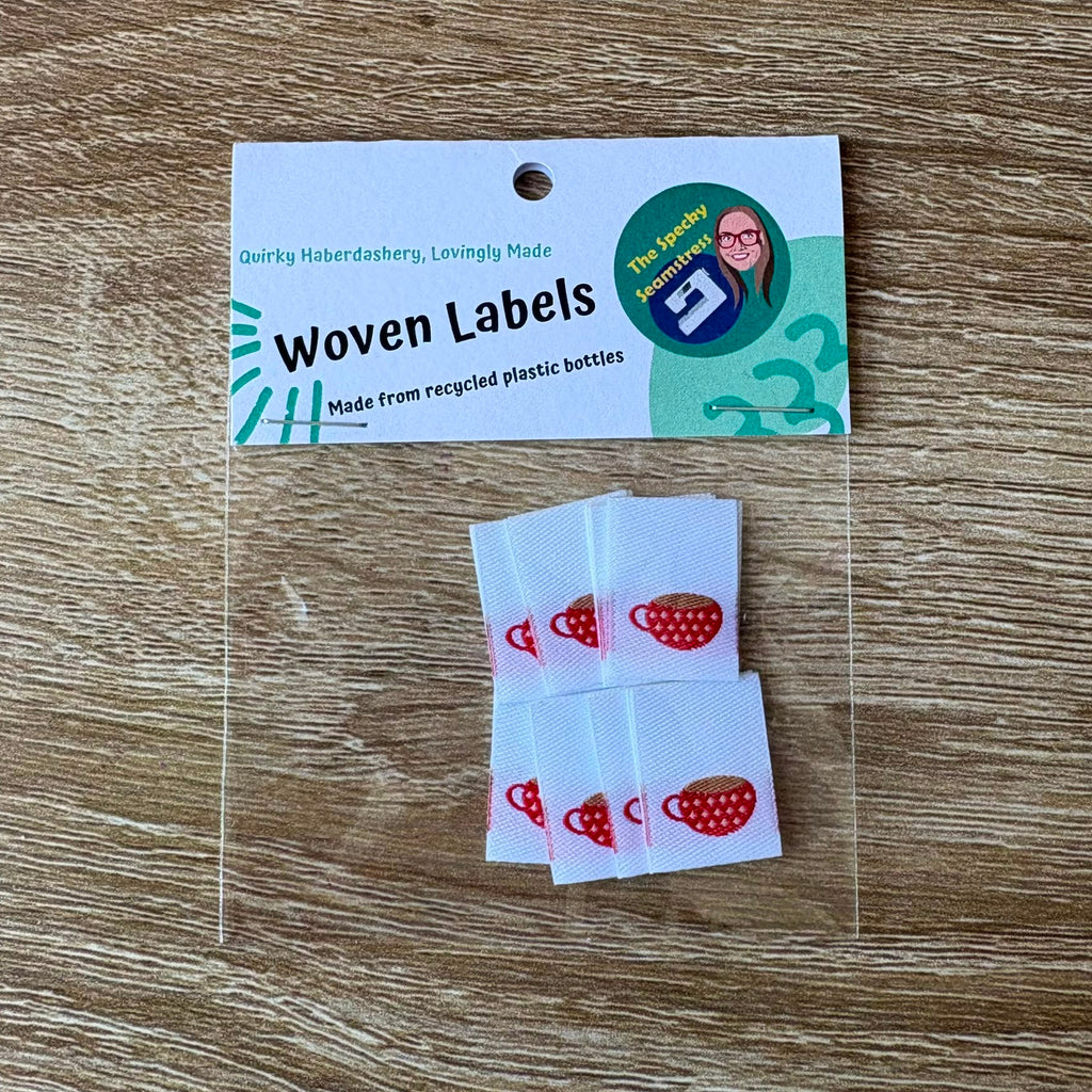 Woven labels with red and white teacup design in a clear packet with a card top, placed on a wooden surface.
