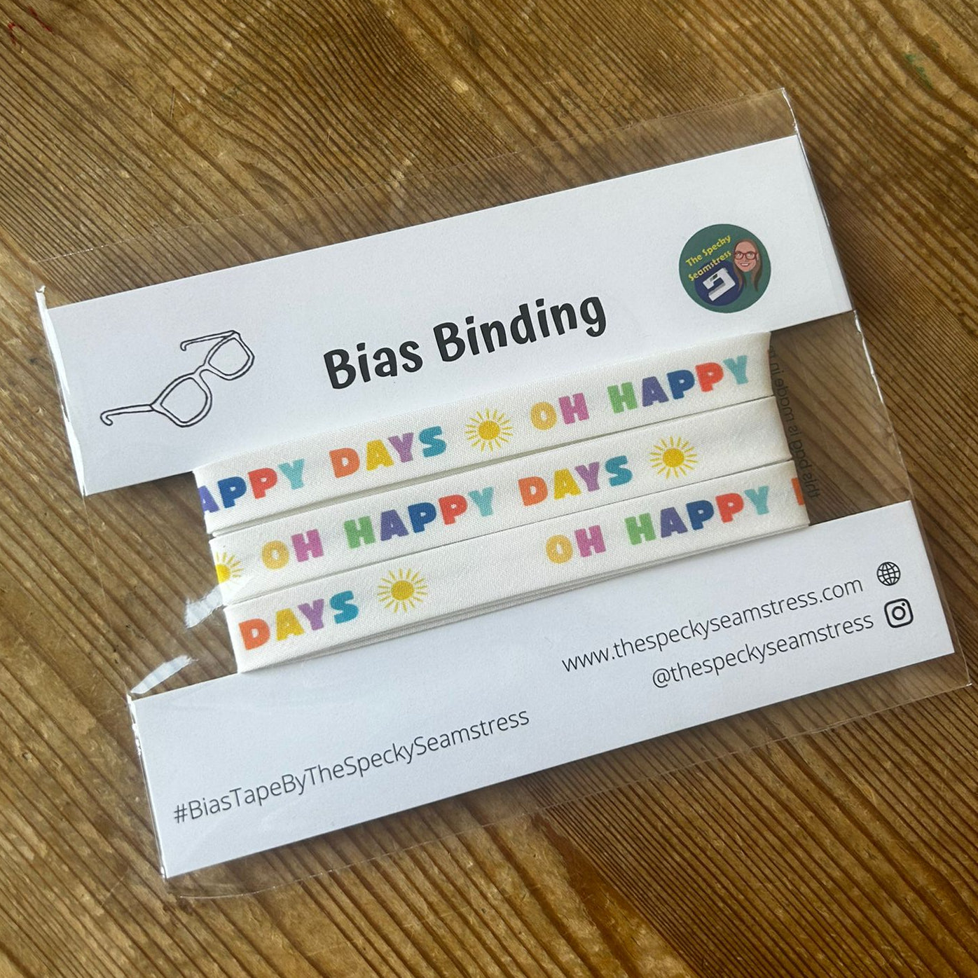 A cream bias tape featuring the words 'Oh Happy Days' in multicolour block font with sunshine motifs, in clear packaging on a white backing card, placed on a wooden surface. 