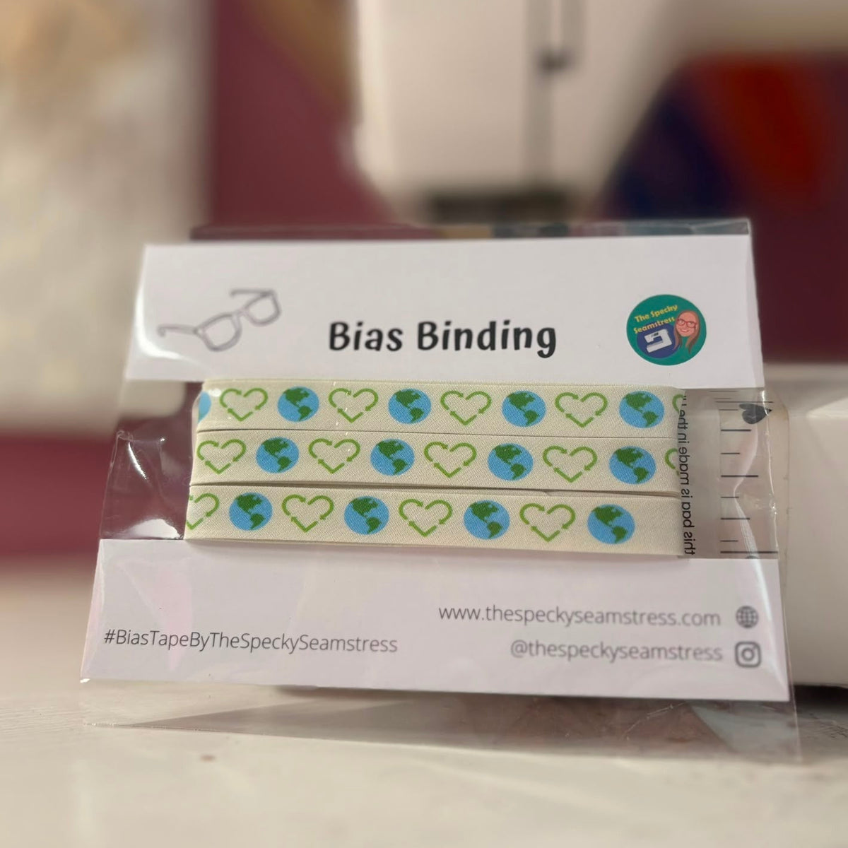 A package of Planet Recycle bias binding propped up against a sewing machine; the design shows planet earth and the recycling symbol in the shape of a heart on a cream background. 