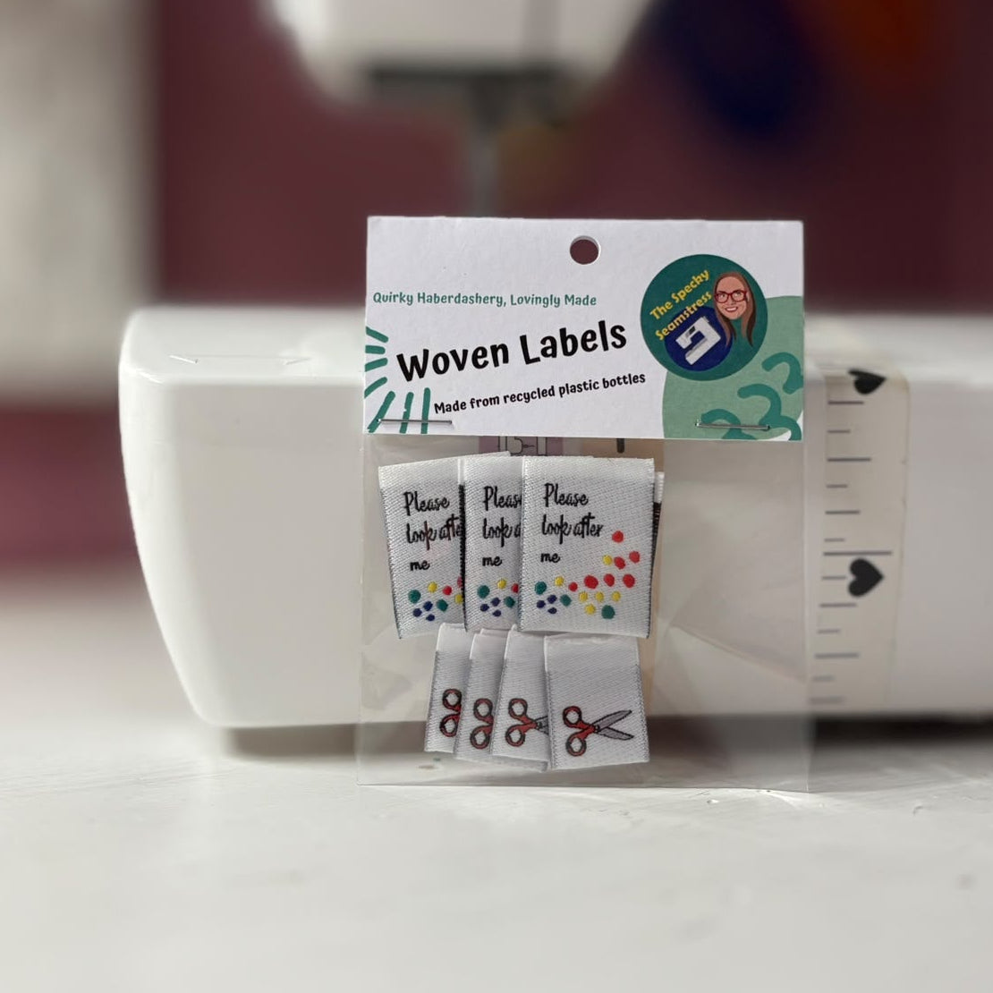 A small packaged set of woven labels in two designs, one reading 'Please Look After Me' with multicolored dots, and the other with mini red handled scissors, displayed on a white sewing machine. The package card says 'Woven Labels' and shows the brand 'The Specky Seamstress'.
