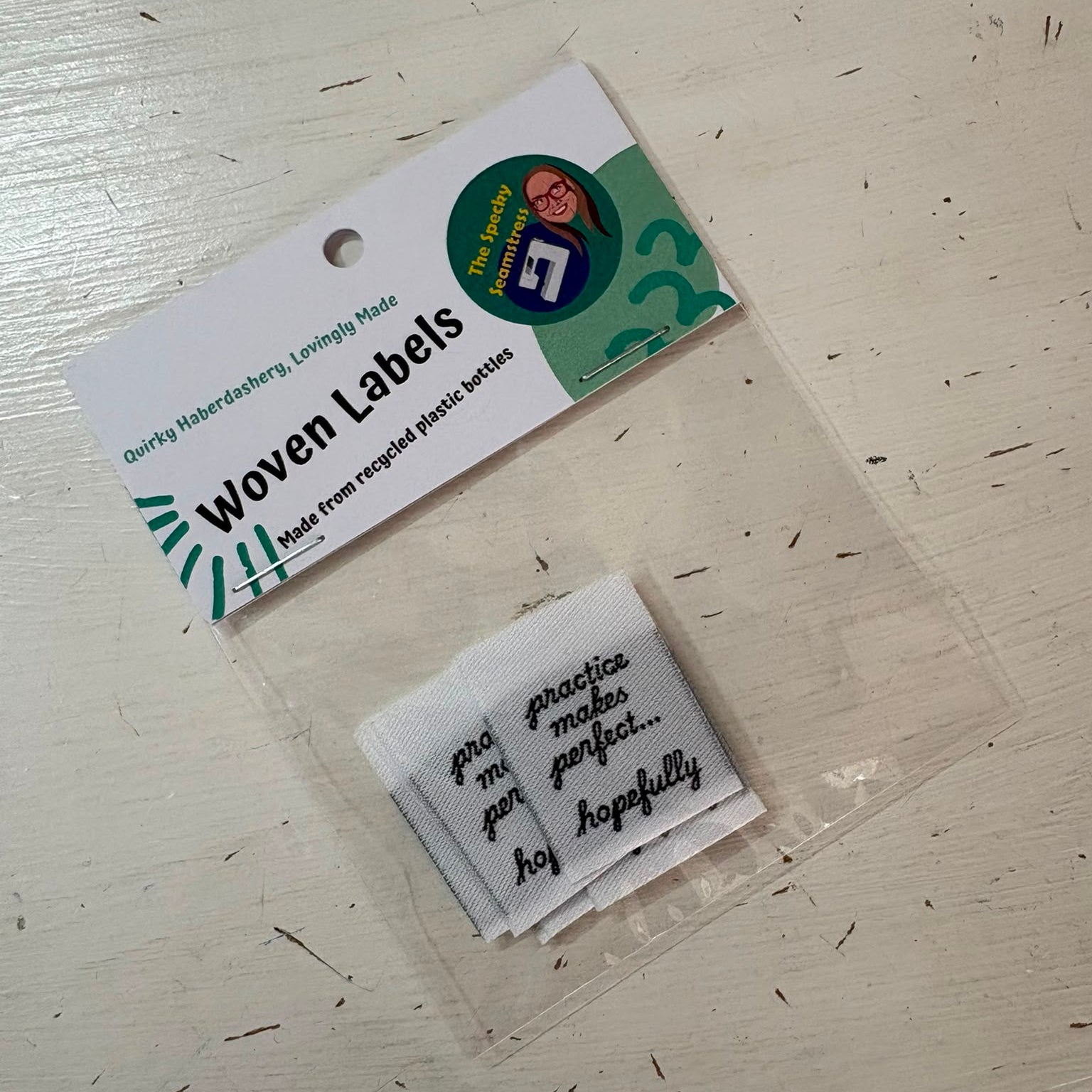 A package of woven labels on a light wood surface. The clear plastic bag contains several small white woven fabric labels with black text, attached to a white paper header card.