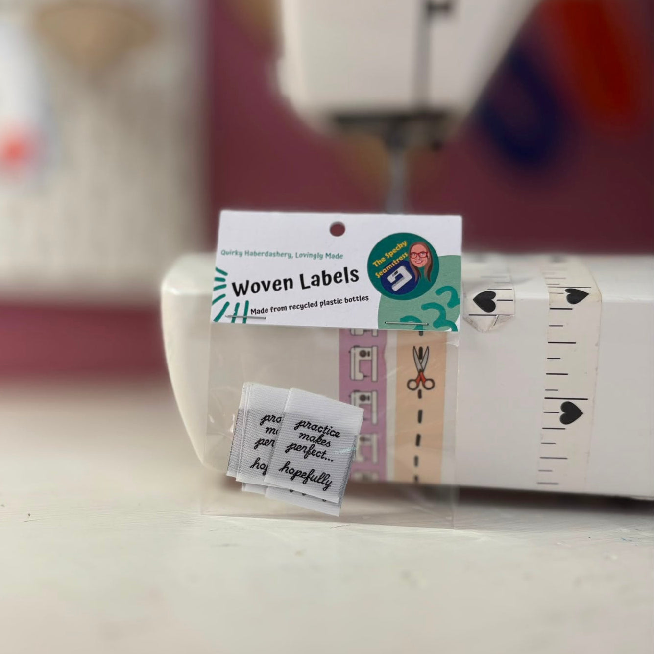 A small clear plastic package labeled 'Woven Labels' containing tiny handwritten fabric labels, photographed in front of a sewing machine on a tabletop.