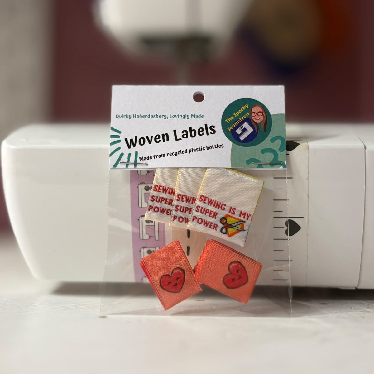 A small clear plastic package of woven labels sits in front of a white sewing machine. The package contains two different designs.