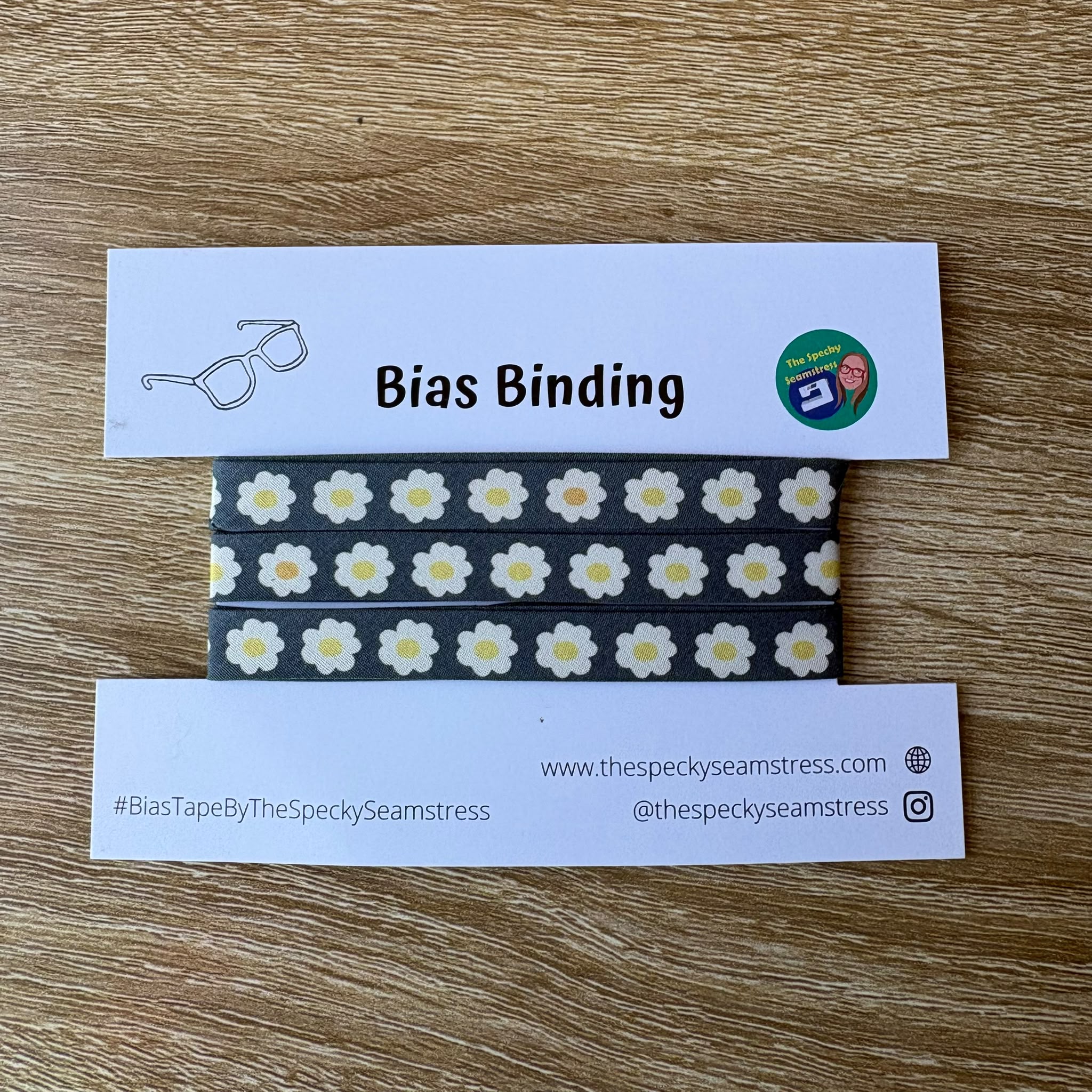 Bias binding tape with daisy pattern on a wooden surface, featuring branding text.