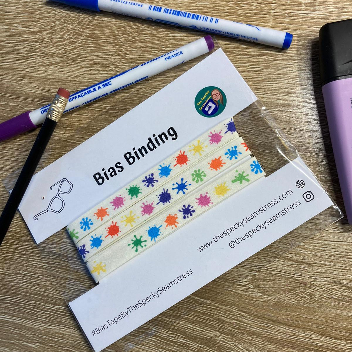 Splat multi-coloured bias binding wrapped on a backing card on a wooden background surrounded by pens and pencils. 