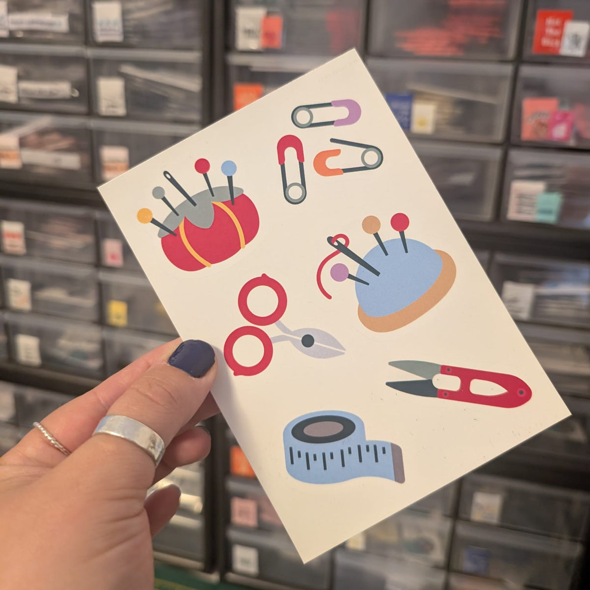 A hand holds a white sticker sheet featuring colorful sewing and crafting tools like scissors, pins, a measuring tape, and a pincushion, with a blurred storage organizer in the background.