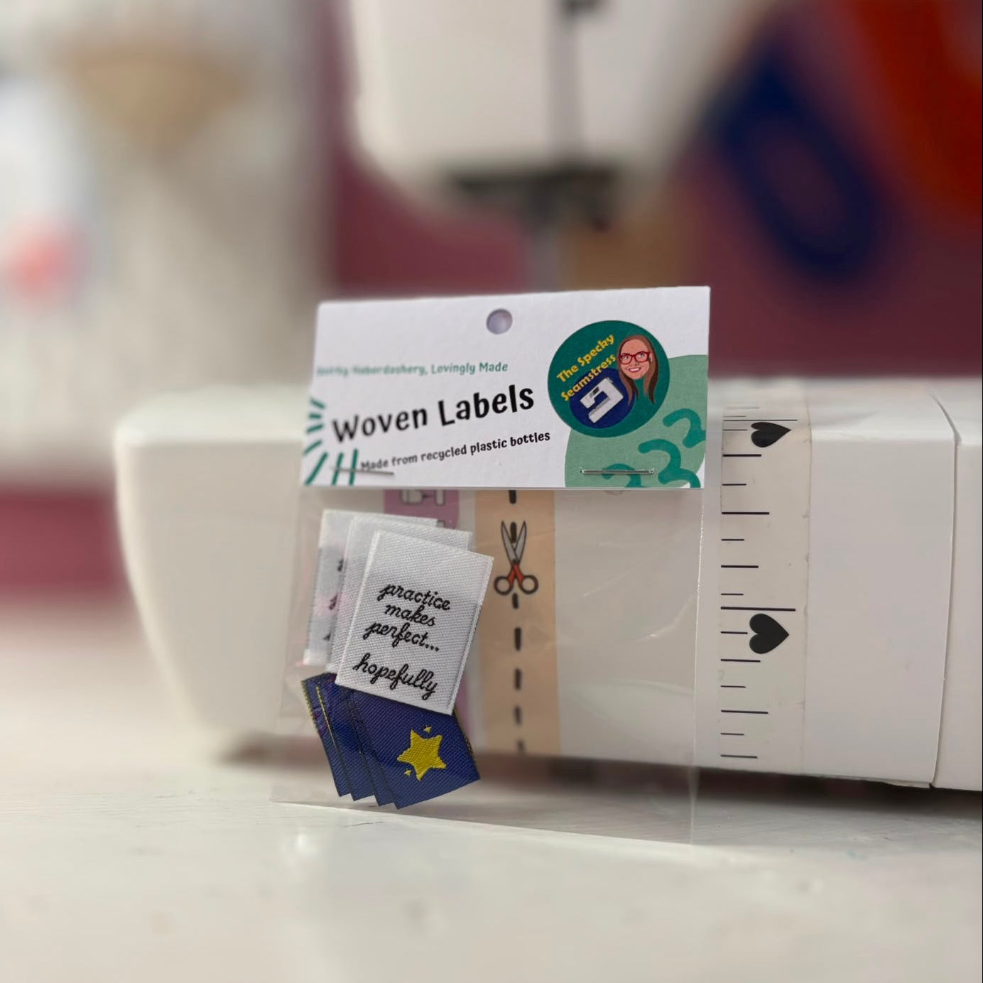 A small clear plastic bag labeled 'Woven Labels' containing several small fabric labels, placed in front of a white sewing machine. Some labels are blue with a yellow star and some are white with black script.