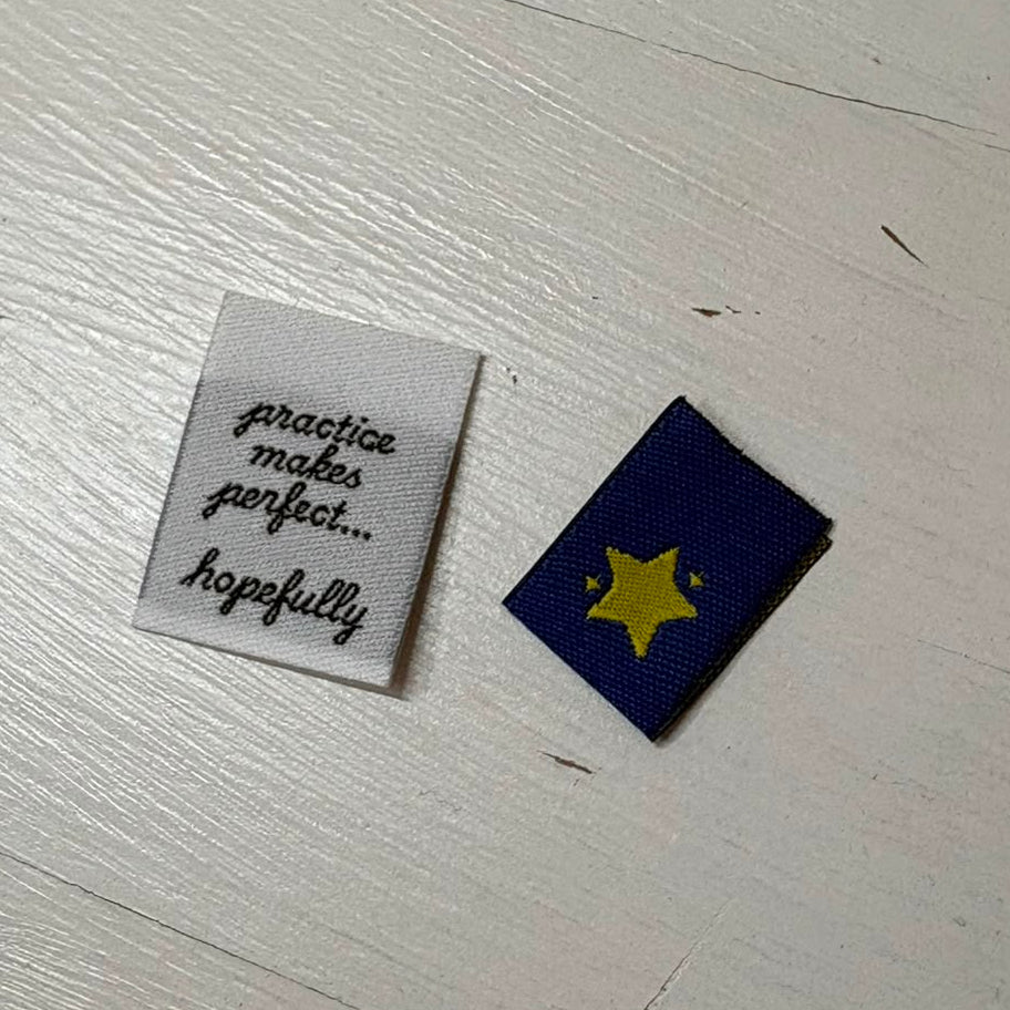 Two small woven labels on a white painted wooden surface. One square label is white with black script text, one is navy with a yellow star.