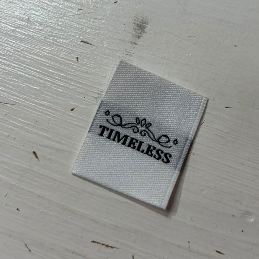 A white woven sewing label on a painted wooden surface; it has the word timeless in a black capitalised font. 