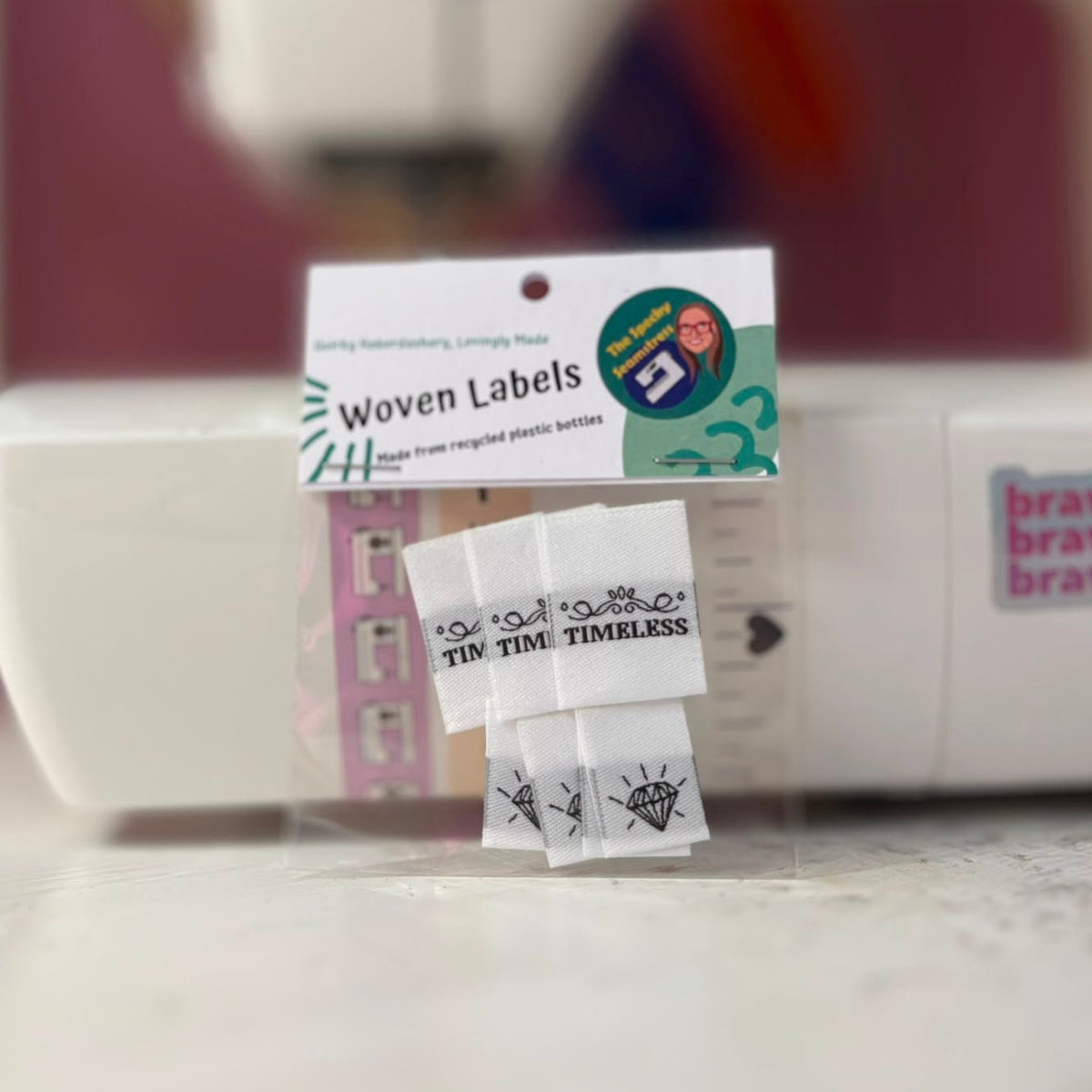 A package of white sewing labels propped up against a sewing machine; the larger label says Timeless in a black capitalised font, the small label shows the outline of a diamond jewel in black.