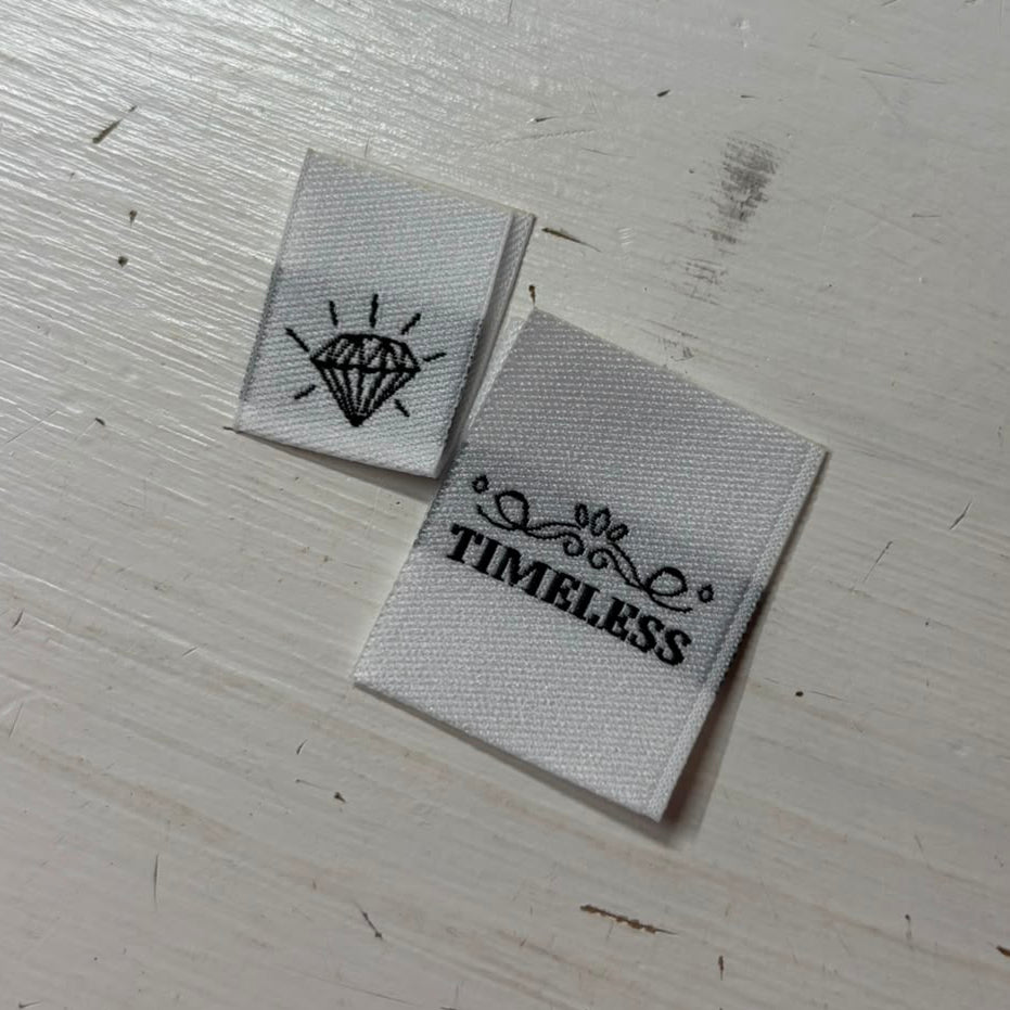 Two white woven sewing labels on a painted wooded surface; one says Timeless in black lettering, one shows a diamond jewel outline. 